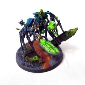 Op de afbeelding: Een groen en blauw geverfd miniatuurbeeldje van een spinachtig wezen met een lang, dun wapen. Het wezen staat op een ronde basis met een zwart en oranje ontwerp.