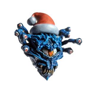 Puede incluir: Un adorno navideño azul y rojo con forma de monstruo con múltiples ojos y un gorro de Papá Noel.