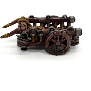 May include: A wooden miniature catapult with a brown and tan paint scheme. The catapult has a rope and a wheel.