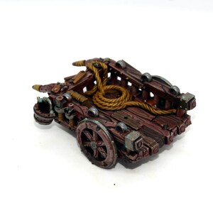May include: A miniature wooden cart with a rope coiled inside. The cart has a brown wooden base with metal accents and a single wheel. The cart is painted in shades of brown, gold, and silver.