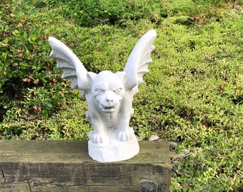 May include: A white gargoyle statue with outstretched wings, perched on a weathered wooden beam. The statue has a detailed face and is surrounded by green foliage and grass, creating a garden or outdoor decor theme.