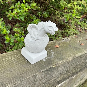 May include: A small, light gray dragon statue emerging from a cracked egg, perched on a square base. The dragon has detailed scales and wings. The statue is made of a stone-like material and is suitable for outdoor garden decor.