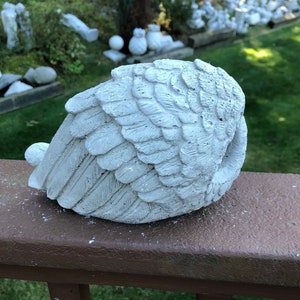 May include: A weathered, white concrete angel wing sculpture. The wing is detailed with individual feather impressions and rests on a brown wooden surface. The sculpture is a decorative garden accent.