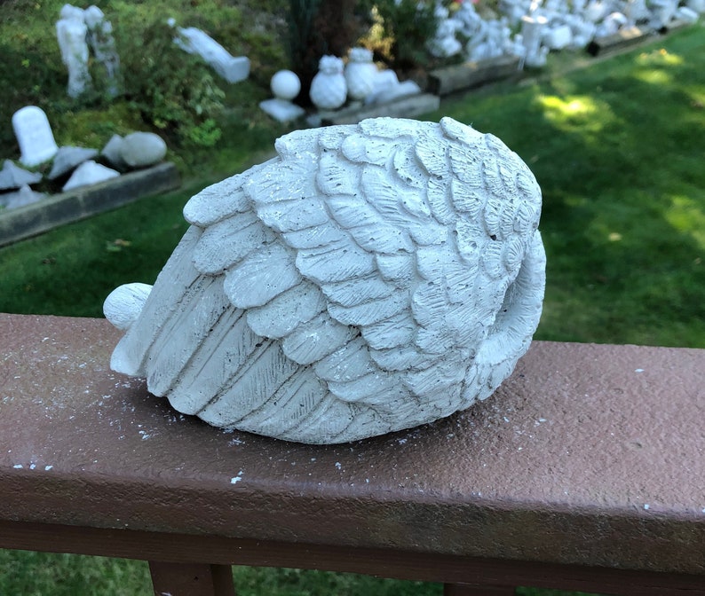 May include: A weathered, white concrete angel wing sculpture. The wing is detailed with individual feather impressions and rests on a brown wooden surface. The sculpture is a decorative garden accent.