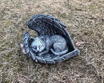 May include: A gray cat sculpture resting peacefully within a pair of angel wings. The cat is curled up, eyes closed, and the wings are intricately detailed. The sculpture is made of a stone-like material and is placed on a bed of dry grass.
