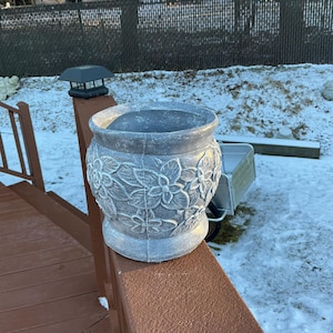 May include: A gray planter with a floral design sits on a wooden railing. The planter is made of a textured material and has a wide, round base.