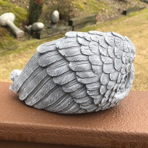 May include: A gray, sculpted angel wing statue with detailed feather textures. The wing is positioned on a brown surface, with a blurred outdoor background. The sculpture is a decorative item, suitable for garden or home decor.