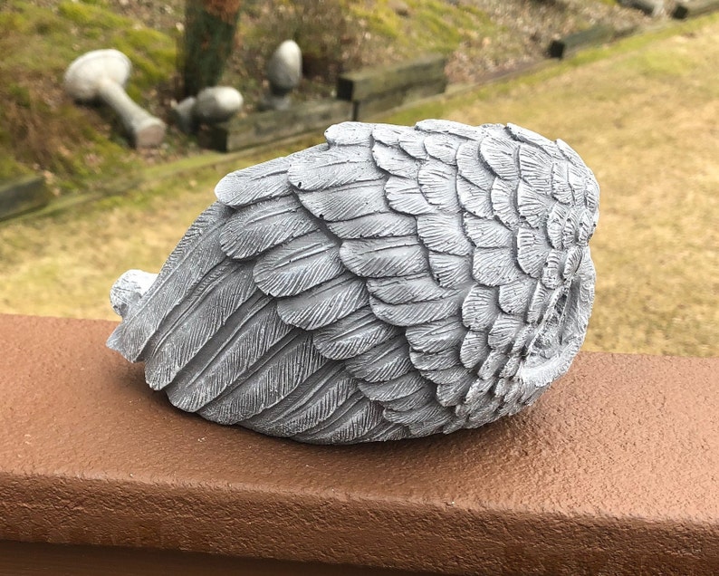 May include: A gray, sculpted angel wing statue with detailed feather textures. The wing is positioned on a brown surface, with a blurred outdoor background. The sculpture is a decorative item, suitable for garden or home decor.