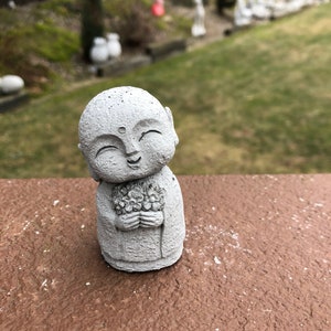 May include: A small, gray concrete statue of a smiling Buddha holding a bouquet of flowers. The statue is sitting on a brown surface.