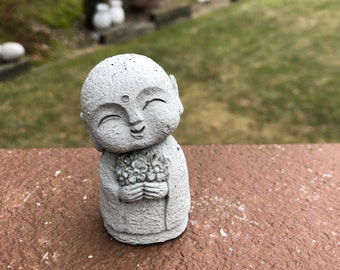 May include: A small, gray concrete statue of a smiling Buddha holding a bouquet of flowers. The statue is sitting on a brown surface.