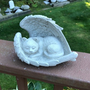 May include: A white, sleeping cat sculpture nestled within angel wings. The cat is curled up, with its eyes closed, and the wings are detailed with feather textures. The sculpture is placed on a brown railing, with a green background.