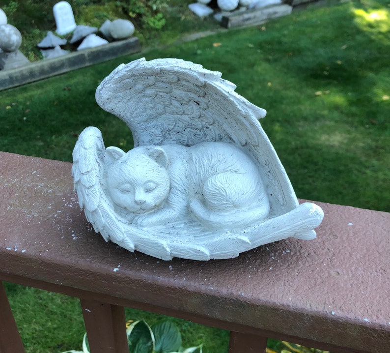 May include: A white, sleeping cat sculpture nestled within angel wings. The cat is curled up, with its eyes closed, and the wings are detailed with feather textures. The sculpture is placed on a brown railing, with a green background.