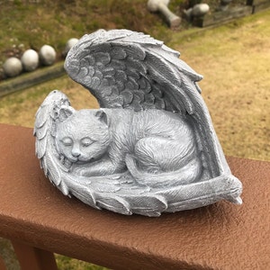 May include: A gray cat sculpture resting peacefully within angel wings. The cat is curled up, appearing asleep, and the wings are detailed with feather textures. The sculpture is made of a stone-like material and is placed on a brown surface.