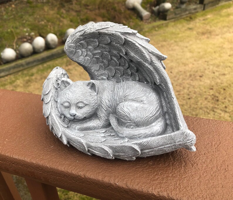 May include: A gray cat sculpture resting peacefully within angel wings. The cat is curled up, appearing asleep, and the wings are detailed with feather textures. The sculpture is made of a stone-like material and is placed on a brown surface.