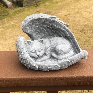 May include: A gray cat sculpture sleeping peacefully within a pair of angel wings. The cat is curled up, with its eyes closed. The wings are detailed with feather textures, creating a serene and comforting scene.