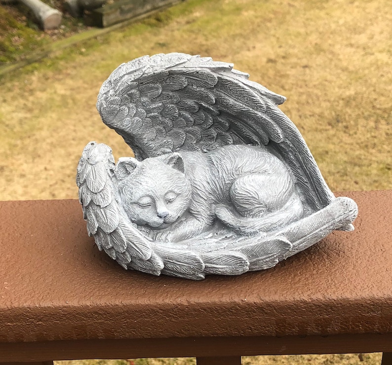 May include: A gray cat sculpture sleeping peacefully within a pair of angel wings. The cat is curled up, with its eyes closed. The wings are detailed with feather textures, creating a serene and comforting scene.