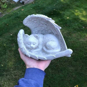 May include: A white cat sculpture sleeping peacefully within a pair of angel wings. The cat is curled up, and the wings are detailed with individual feathers. The sculpture is made of a light-colored material, possibly concrete or stone.