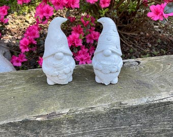 May include: Two white concrete garden gnomes with pointed hats and holding objects. The gnomes are on a weathered wooden surface, with vibrant pink flowers in the background. The gnomes are detailed with textured clothing and facial features.