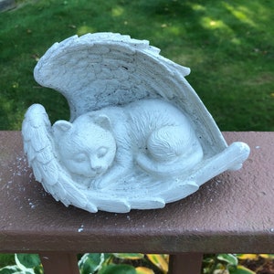 May include: A white cat figurine sleeping peacefully within a pair of angel wings. The sculpture is made of a light-colored material and rests on a brown surface. The cat is curled up, with its eyes closed, and the wings are detailed with feather textures.