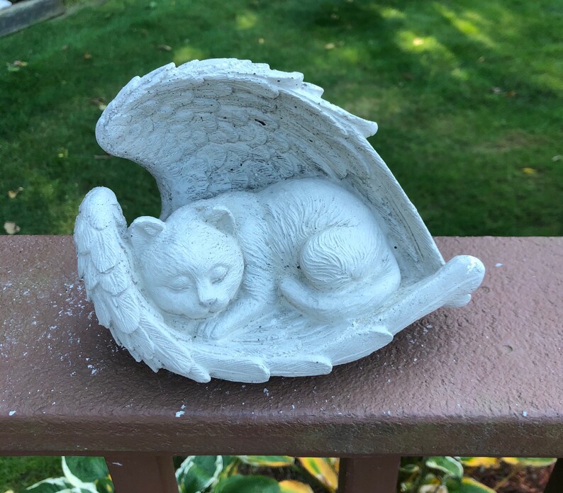 May include: A white cat figurine sleeping peacefully within a pair of angel wings. The sculpture is made of a light-colored material and rests on a brown surface. The cat is curled up, with its eyes closed, and the wings are detailed with feather textures.