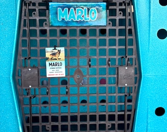 May include: A bright teal RuffLand Performance Kennel crate featuring a black grid door. A rectangular sign with the name MARLO in blue letters is attached. Below it, a tag displays a German Shepherd photo and the name MARLO.