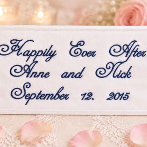 May include: A rectangular white fabric sign featuring dark blue embroidered text that reads Happily Ever After Anne and Nick September 12. 2015. The sign has a stitched border and is displayed in a horizontal orientation with a clean finish.