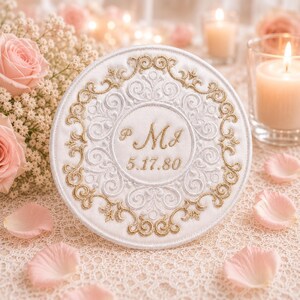 May include: A circular white fabric ornament featuring intricate gold and white embroidery in a scrollwork pattern. The center displays the embroidered initials PMJ and the date 5.17.80 in gold thread, surrounded by a decorative gold border.
