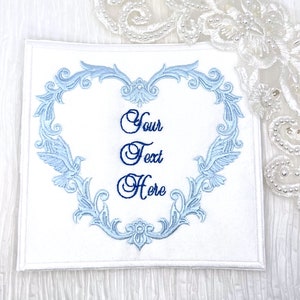 May include: White fabric square with a blue embroidered heart design. The heart is surrounded by a decorative floral pattern and two birds. The text "Your Text Here" is embroidered in the center of the heart.