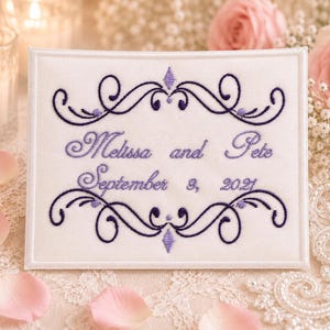May include: A rectangular white fabric sign featuring purple machine embroidery. The text reads "Melissa and Pete" and "September 3, 2021" centered between two decorative purple scrollwork flourishes with small diamond-shaped accents.
