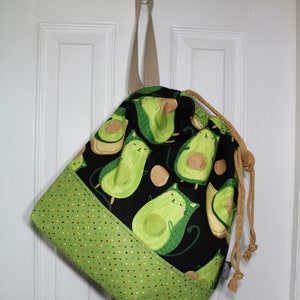 Puede incluir: Una bolsa de cord&oacute;n negro con un estampado de aguacate verde y marr&oacute;n. La bolsa tiene un fondo de lunares verde y marr&oacute;n y un cord&oacute;n de color marr&oacute;n.