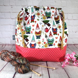 May include: A drawstring knitting project bag with a white background featuring garden gnomes, flowers, and gardening tools. The bottom of the bag is red with white polka dots. Yarn and knitting needles are in the foreground.