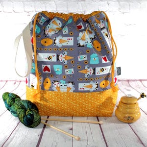 May include: A gray drawstring project bag with a bee and ice cream cone pattern, a yellow honeycomb base, and a cream-colored handle. The bag is next to yarn, knitting needles, and a honey pot.