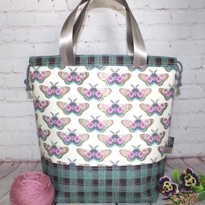 May include: A tote bag featuring a repeating pattern of moths with pink, teal, and yellow wings on a cream background. The bag has a dark teal and black plaid base, two grey nylon handles, and a drawstring closure at the top with grey cords.