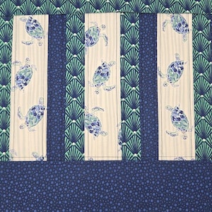 May include: A fabric with a repeating pattern of sea turtles in blue and white on a cream background. The design is divided into vertical stripes with a blue and green fan-like pattern on the sides. The bottom has a blue dotted pattern.