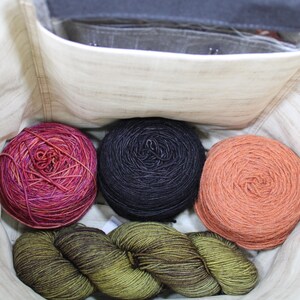 May include: A cream-colored fabric bag containing three round balls of yarn in variegated magenta, solid black, and solid peach, along with a single twisted skein of olive green yarn resting at the bottom of the interior compartment.