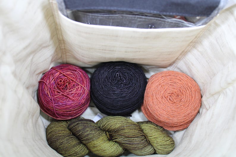 May include: A cream-colored fabric bag containing three round balls of yarn in variegated magenta, solid black, and solid peach, along with a single twisted skein of olive green yarn resting at the bottom of the interior compartment.