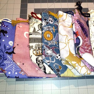 May include: A collection of eight fabric drawstring bags with various patterns. The bags feature designs such as bats, skulls, spiderwebs, and celestial motifs. The color palette includes purple, pink, and cream, with black and gold accents. Each bag has a drawstring closure.