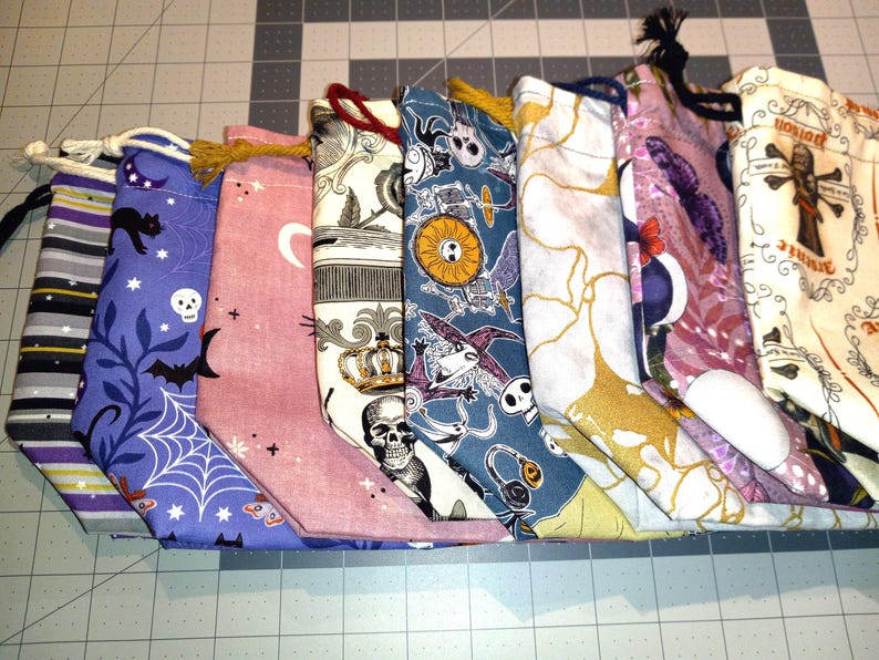 May include: A collection of eight fabric drawstring bags with various patterns. The bags feature designs such as bats, skulls, spiderwebs, and celestial motifs. The color palette includes purple, pink, and cream, with black and gold accents. Each bag has a drawstring closure.