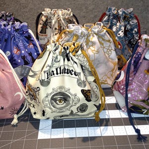 May include: A collection of drawstring bags in various patterns and colors. Designs include celestial motifs, Halloween-themed graphics with the word "Halloween", and floral prints. Each bag has a drawstring closure.