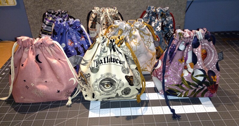 May include: A collection of drawstring bags in various patterns and colors. Designs include celestial motifs, Halloween-themed graphics with the word "Halloween", and floral prints. Each bag has a drawstring closure.