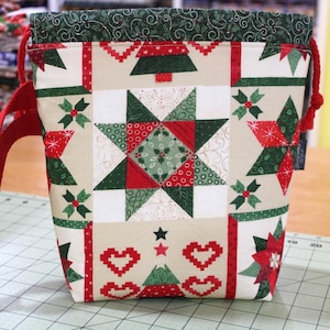 May include: A fabric drawstring bag featuring a festive holiday print with red and green stars, Christmas trees, and heart motifs on a cream background. The bag has a dark green top border with a subtle pattern and red cord handles on each side.