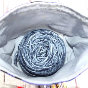 May include: A gray and blue yarn ball inside a white and purple lined fabric bag. The yarn is wound tightly and secured with a clear band. The bag is open, revealing the yarn inside.