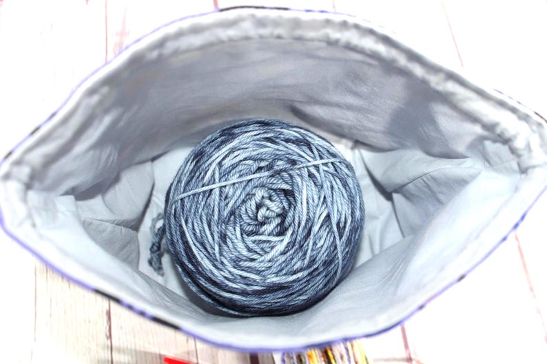 May include: A gray and blue yarn ball inside a white and purple lined fabric bag. The yarn is wound tightly and secured with a clear band. The bag is open, revealing the yarn inside.