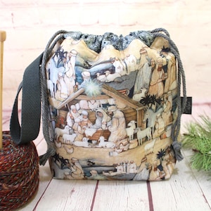 Puede incluir: Una bolsa de tela con cordón ajustable que presenta una escena de natividad impresa con figuras, animales y un establo. La bolsa tiene una correa gris oscuro y cierres de cordón gris. Se muestra junto a un ovillo de lana y dos agujas de tejer de madera.