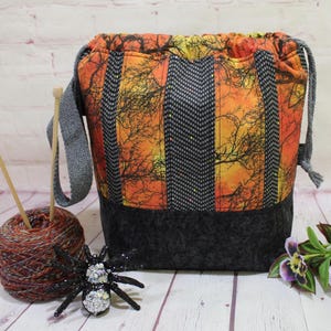 May include: A fabric drawstring yarn bag with a colourful autumn tree design. The bag features a grey handle and base, with a matching drawstring. A ball of yarn, knitting needles, and a decorative spider are next to the bag.