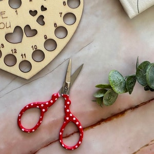 May include: Red and white polka dot scissors with a silver blade. The scissors are on a white surface with a wooden heart-shaped craft project in the background.
