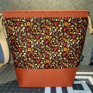 May include: A drawstring bag featuring a dark brown central panel with a repeating pattern of autumn leaves, mushrooms, and acorns. The top and bottom sections are made of orange fabric with a small white cross pattern. Includes beige rope ties.