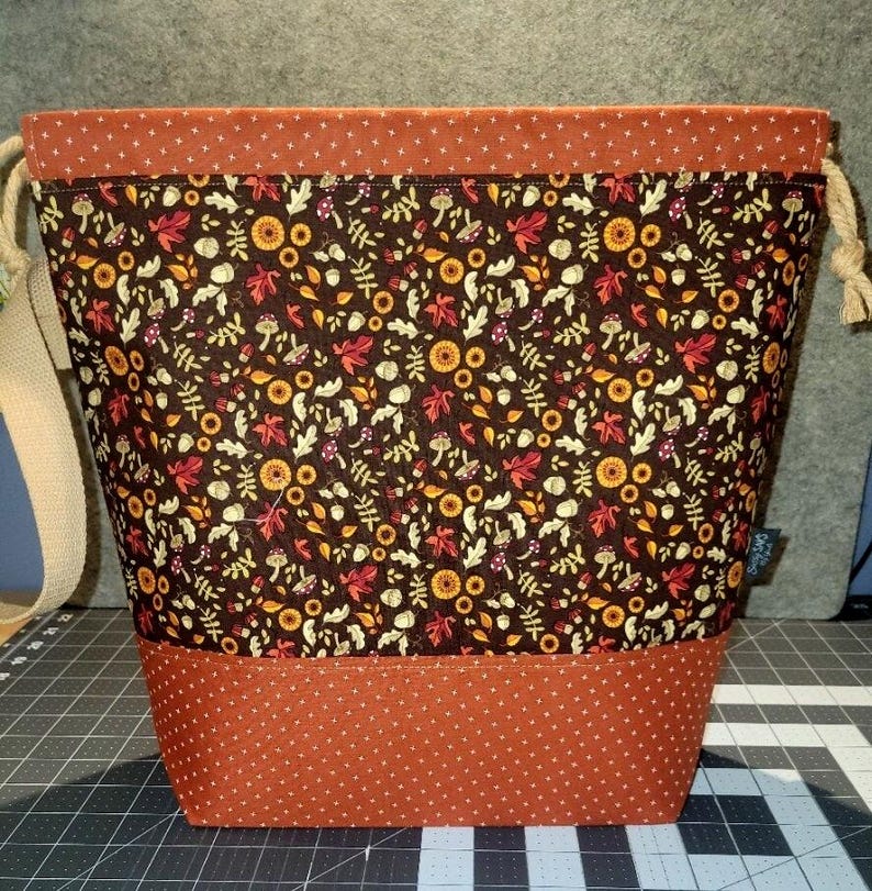 May include: A drawstring bag featuring a dark brown central panel with a repeating pattern of autumn leaves, mushrooms, and acorns. The top and bottom sections are made of orange fabric with a small white cross pattern. Includes beige rope ties.
