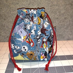 May include: A drawstring pouch with a teal background featuring a pattern of characters from The Nightmare Before Christmas. The bag has a red drawstring closure. The pouch is made of fabric.
