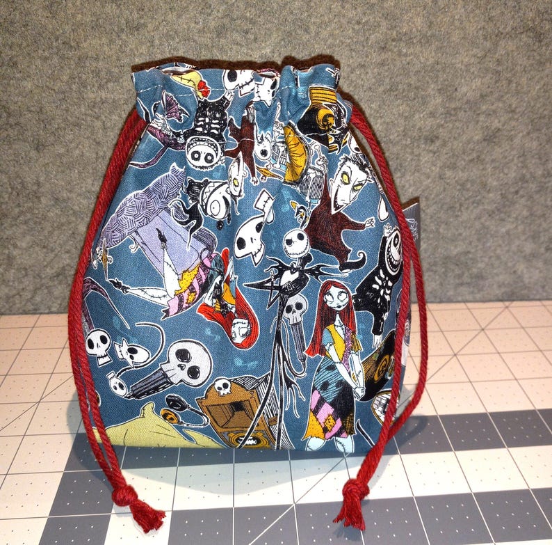 May include: A drawstring pouch with a teal background featuring a pattern of characters from The Nightmare Before Christmas. The bag has a red drawstring closure. The pouch is made of fabric.